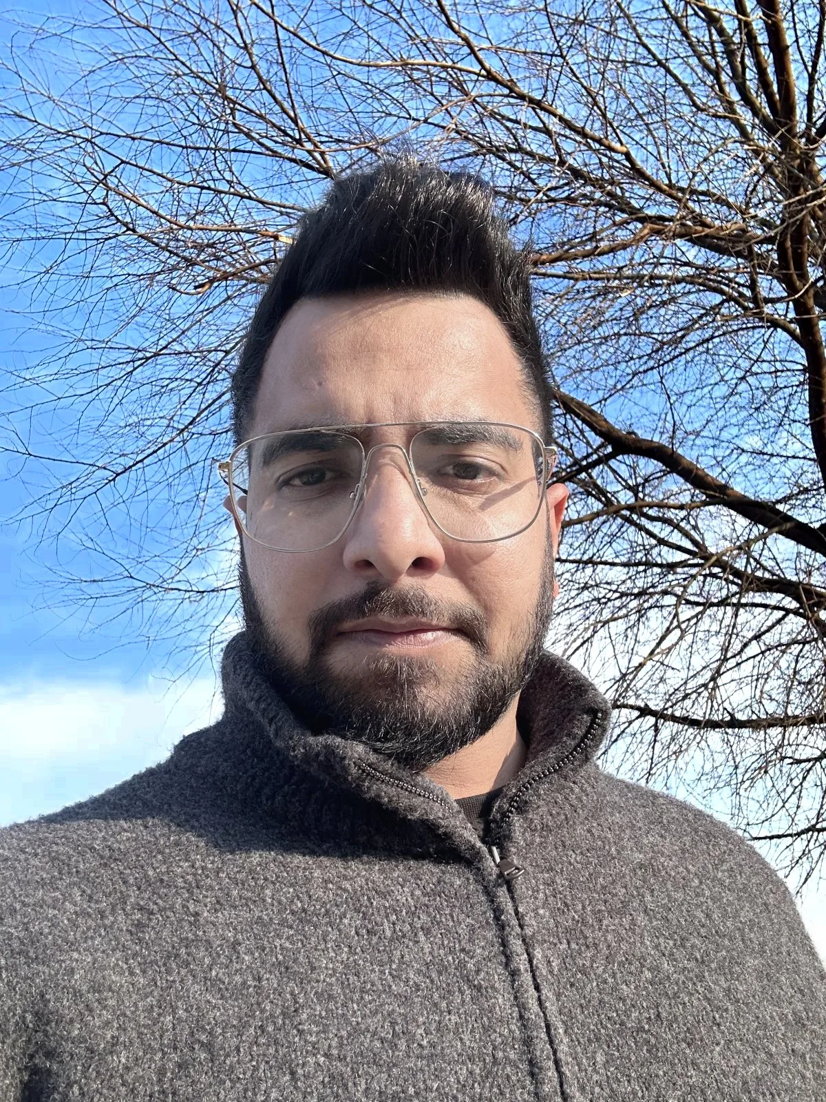 Close-up of Salman Ansari in glasses and a grey knit zip jumper, under bare winter branches and a blue sky